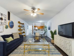 Apartments in Fayetteville, North Carolina Living room with a blue sofa, glass coffee table, TV on a white console, ceiling fan, wall art, and wooden shelving. Dining area and kitchen are visible in the background. Stone Ridge Apartments in Fayetteville 3001 Stone Carriage Circle Fayetteville, NC 28304