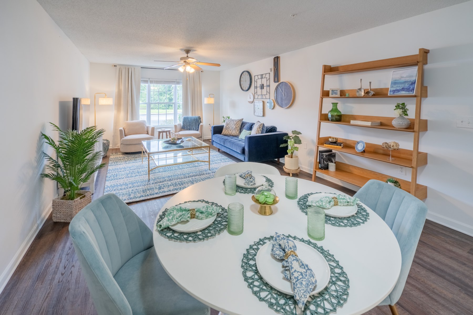 Apartments in Fayetteville, North Carolina Step into the modern open-plan living room and dining area at Stone Ridge Apartments, featuring a round table, blue chairs, decorative shelving, and a cozy blue sofa. A ceiling fan complements the bright window and lush greenery that enhance this inviting space. Stone Ridge Apartments in Fayetteville 3001 Stone Carriage Circle Fayetteville, NC 28304