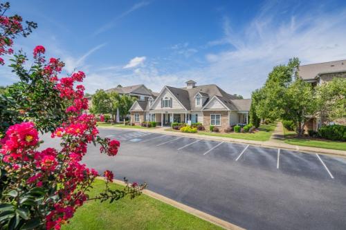 Apartments in Fayetteville, North Carolina Suburban neighborhood with a stone house, parking lot, manicured lawns, and flowering trees under a blue sky. Stone Ridge Apartments in Fayetteville 3001 Stone Carriage Circle Fayetteville, NC 28304