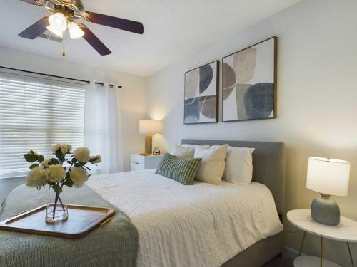 Apartments in Fayetteville, North Carolina A bedroom with a bed, two side tables with lamps, a ceiling fan, and abstract art on the wall. A vase of flowers and a wooden tray are on the bed.