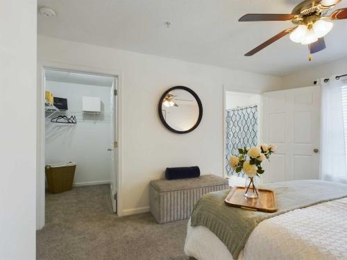 Apartments in Fayetteville, North Carolina A bedroom with a ceiling fan, an attached closet, and a bed adorned with a gray blanket and a decorative bouquet. A round mirror and a cushioned bench are on the wall adjacent to the closet.