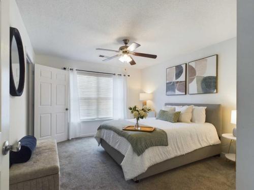 Apartments in Fayetteville, North Carolina A bedroom with a gray bed, white bedding, and green pillows. The room has a ceiling fan, a window with white curtains, and two abstract paintings above the bed. There is a white nightstand with a lamp.