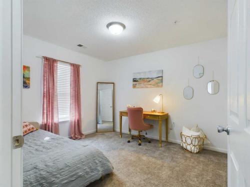 Apartments in Fayetteville, North Carolina A bedroom with white walls features a bed, a window with pink curtains, a full-length mirror, a desk with a chair and lamp, wall art, and round mirrors above a basket with linens.
