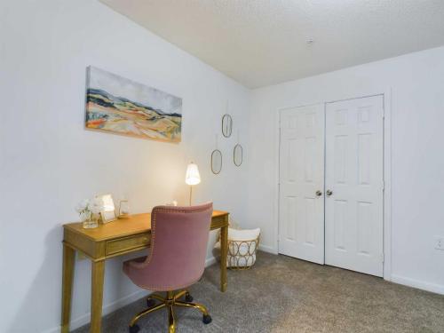 Apartments in Fayetteville, North Carolina A home office with a pink chair at a wooden desk, a painting above, and mirrors on the wall. A lamp, plants, and decor items are on the desk. Two closed double closet doors are in the background.