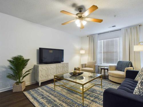 Apartments in Fayetteville, North Carolina A living room with a ceiling fan, TV on a console, a glass coffee table, a patterned rug, potted plant, two chairs, a couch, and a floor lamp in front of a window with blinds and curtains.