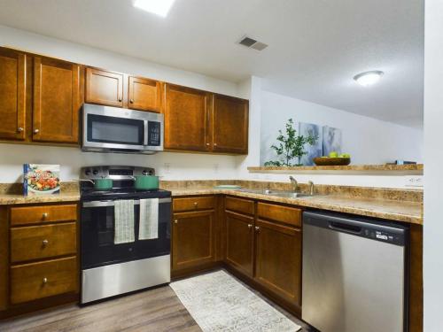 Apartments in Fayetteville, North Carolina A modern kitchen with brown cabinets, stainless steel appliances including a microwave, stove, dishwasher, and a countertop with a cookbook displayed. There is a small potted plant and a bowl of green apples.