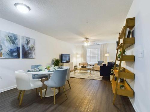 Apartments in Fayetteville, North Carolina A modern living and dining area with a round table set for four, a mounted TV, a seating area with a sofa and chairs, and a wooden bookshelf on the right. The room features wooden flooring and contemporary decor.