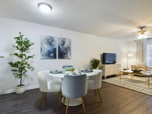 Apartments in Fayetteville, North Carolina A modern living and dining area with a round table set for four, abstract wall art, potted plants, a TV on a console, and a rug on dark wood flooring.