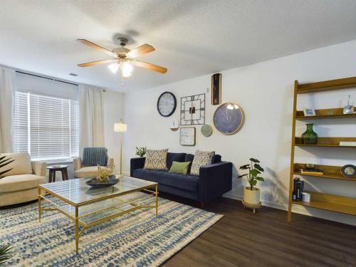 Apartments in Fayetteville, North Carolina A modern living room with a blue sofa, two beige chairs, a glass coffee table, a wall with clocks and decor, a ceiling fan, a large window with curtains, and a wooden shelf with various items.