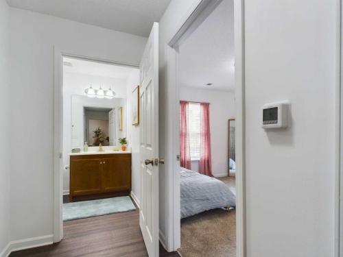 Apartments in Fayetteville, North Carolina A view of a hallway with doors to a bathroom and a bedroom. The bathroom features a vanity with dual sinks, and the bedroom has a bed with a blue quilt and pink curtains. A thermostat is on the wall.