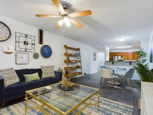 Apartments in Fayetteville, North Carolina Modern living room with dark blue sofa, gold coffee table, and various wall clocks. Ceiling fan above and dining area with a wooden table and white chairs in the background. Decor includes plants and shelves.